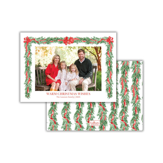 A family portrait of a man, woman, and two children sitting together on a bench, framed by a wreath of greenery and red ribbons.