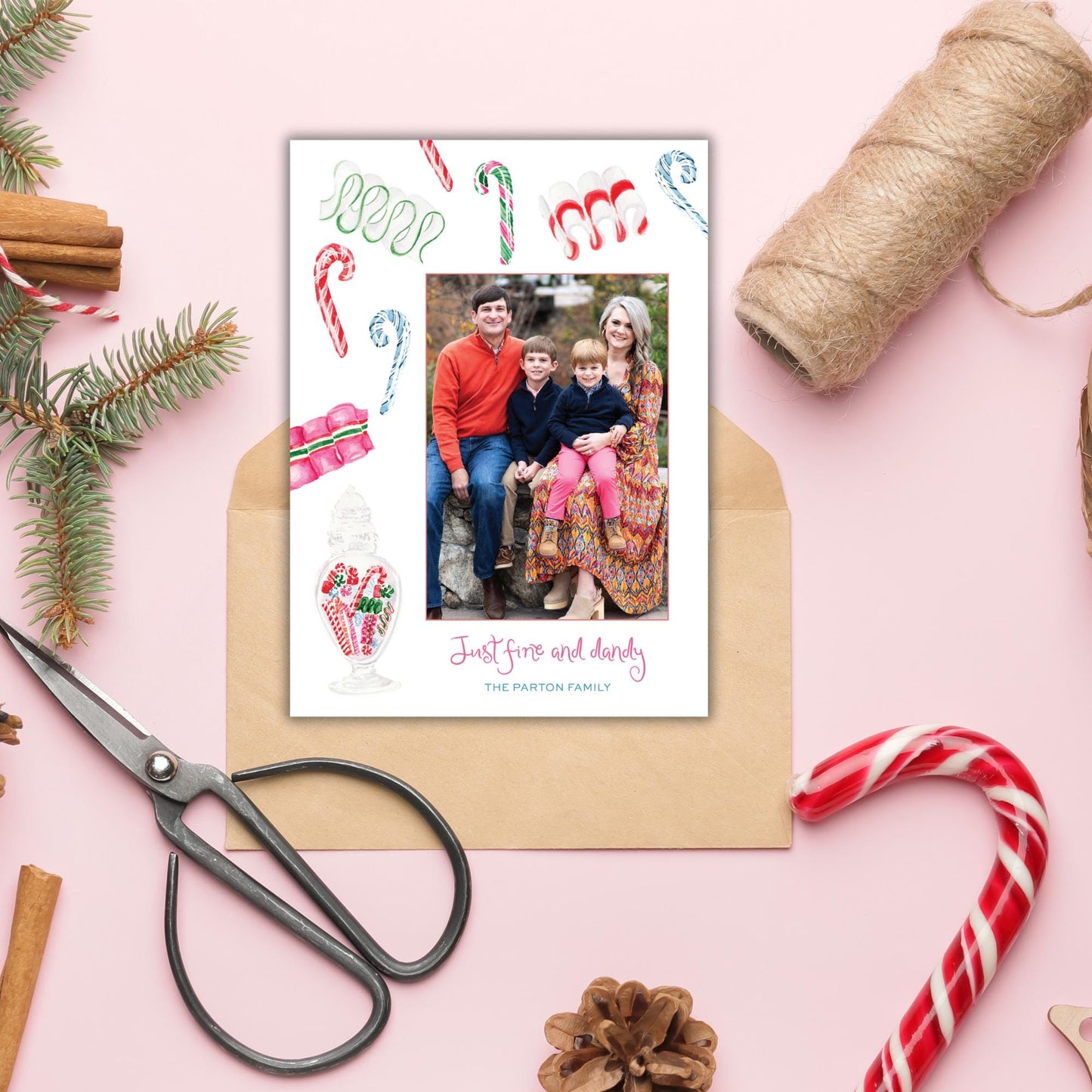 A family portrait is displayed on a pink background, surrounded by holiday decorations such as candy canes, pine branches, and a pair of scissors.