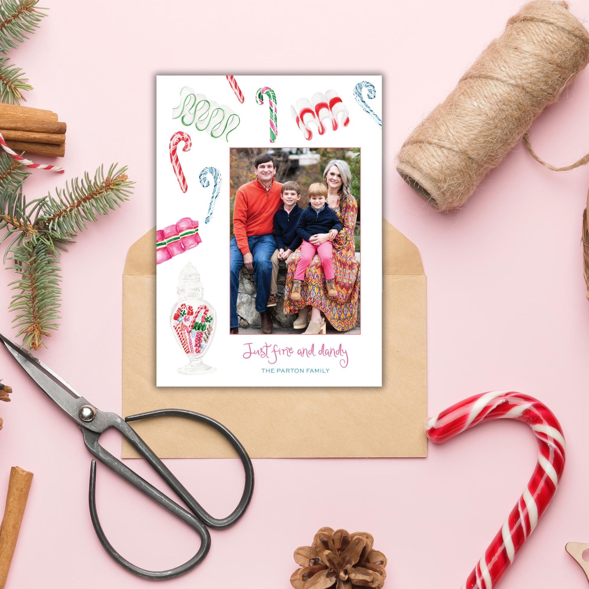 A family portrait is displayed on a pink background, surrounded by holiday decorations such as candy canes, pine branches, and a pair of scissors.