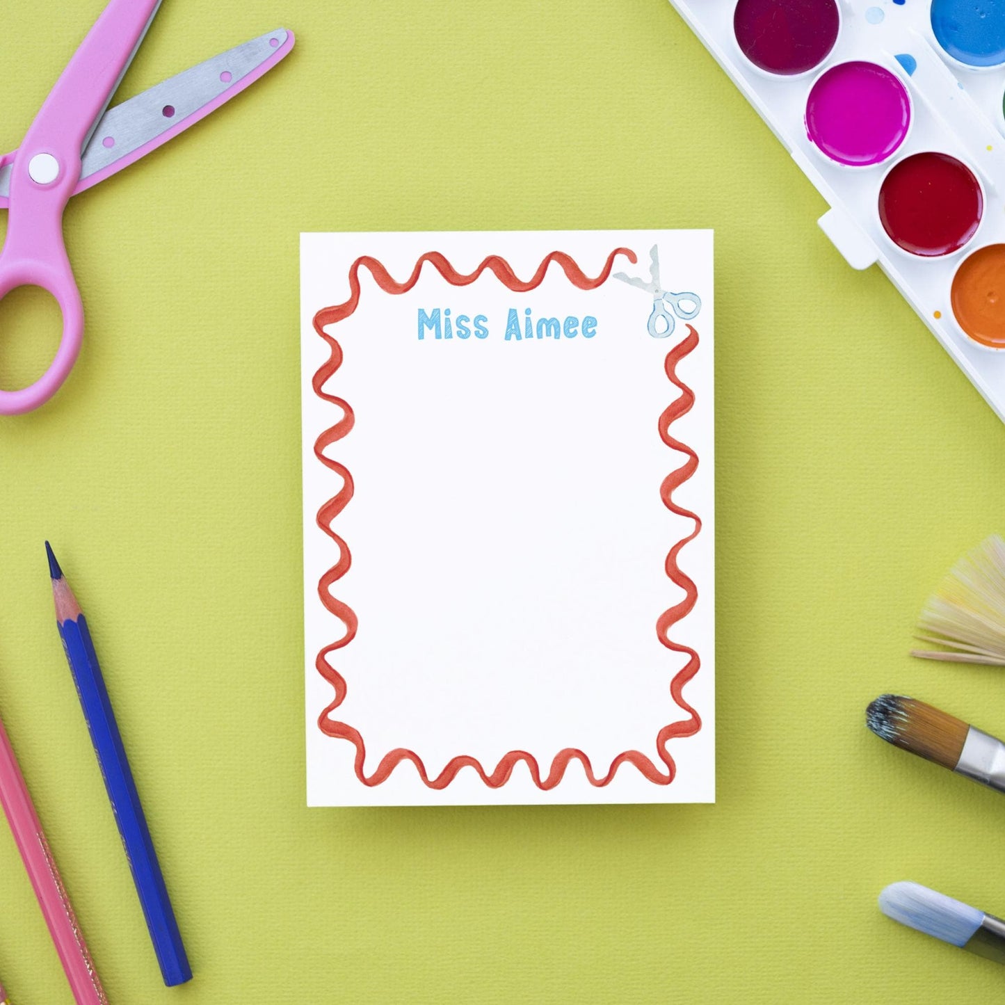 a blank white notepad with a red wavy border, surrounded by various art supplies such as scissors, paintbrushes, and colored pencils on a green background.