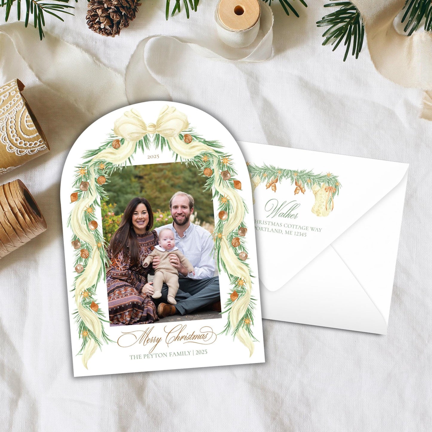 A Christmas-themed card with a family photo of a man, woman, and child, surrounded by a decorative wreath and pine cones. The card is placed on a white cloth, and there is an envelope visible in the background.