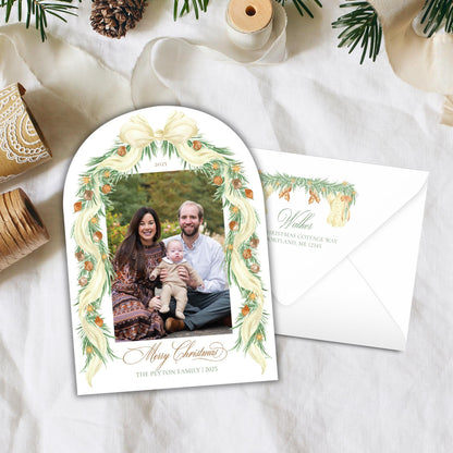 A Christmas-themed card with a family photo of a man, woman, and child, surrounded by a decorative wreath and pine cones. The card is placed on a white cloth, and there is an envelope visible in the background.