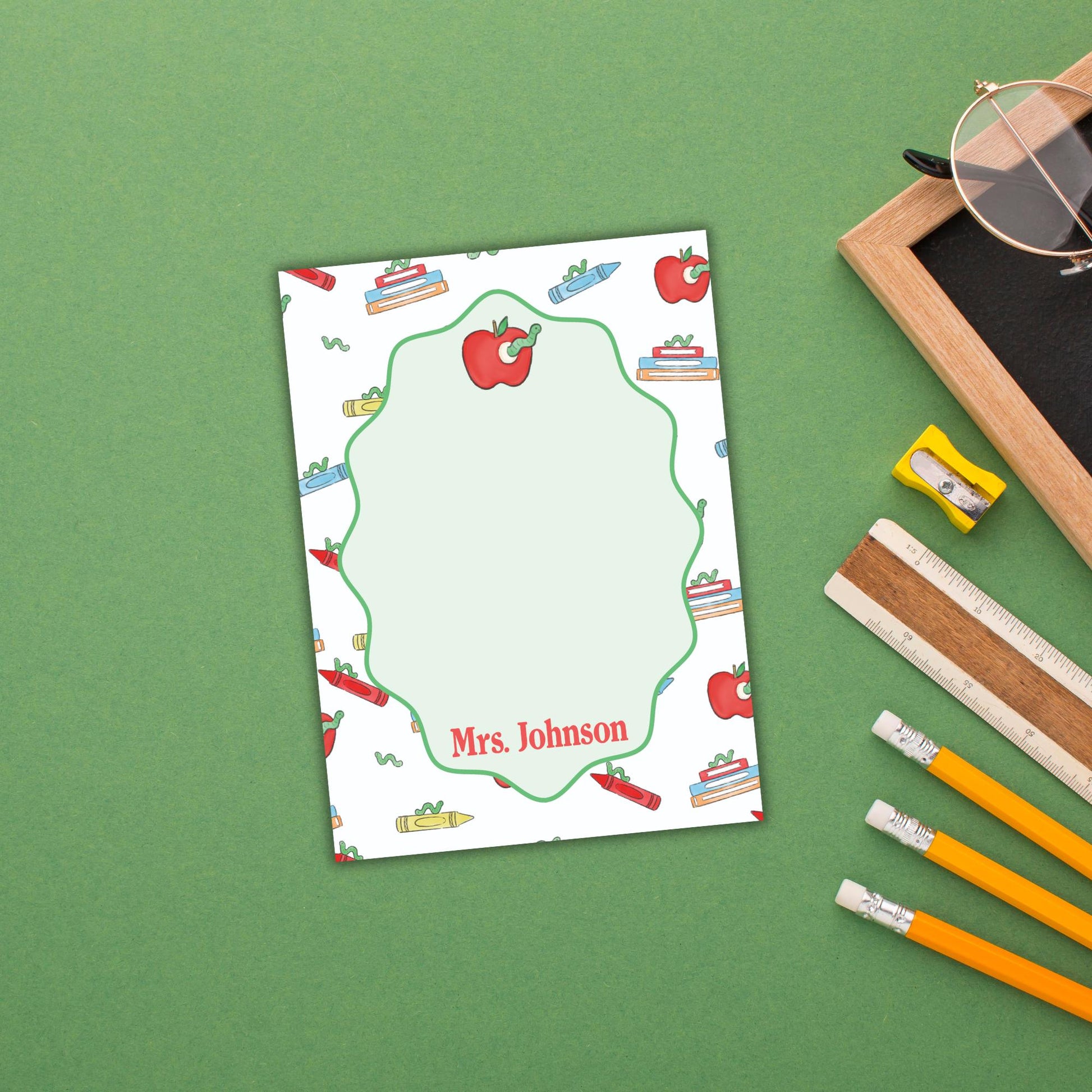 A personalized nameplate with a green border and a pattern of apples and books, placed on a green surface next to a pair of glasses and a ruler.