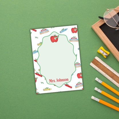 A personalized nameplate with a green border and a pattern of apples and books, placed on a green surface next to a pair of glasses and a ruler.