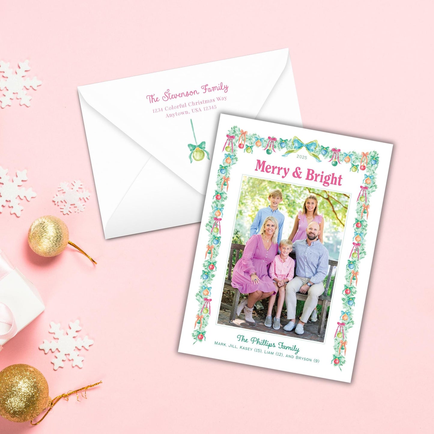 A white envelope with a holiday-themed card featuring a family photo, surrounded by snowflakes and ornaments on a pink background.