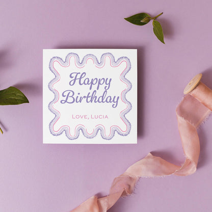 A white greeting card with a pink and purple wavy border, placed on a purple background. The card has the text "Happy Birthday" written on it, and there is a pink ribbon and some leaves nearby.