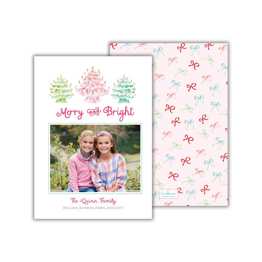 A Christmas card featuring a photo of two children, likely siblings, sitting together in a garden setting. The card has a pattern of red and green bows on the background, and the text &quot;Merry and Bright&quot; is written above the photo.