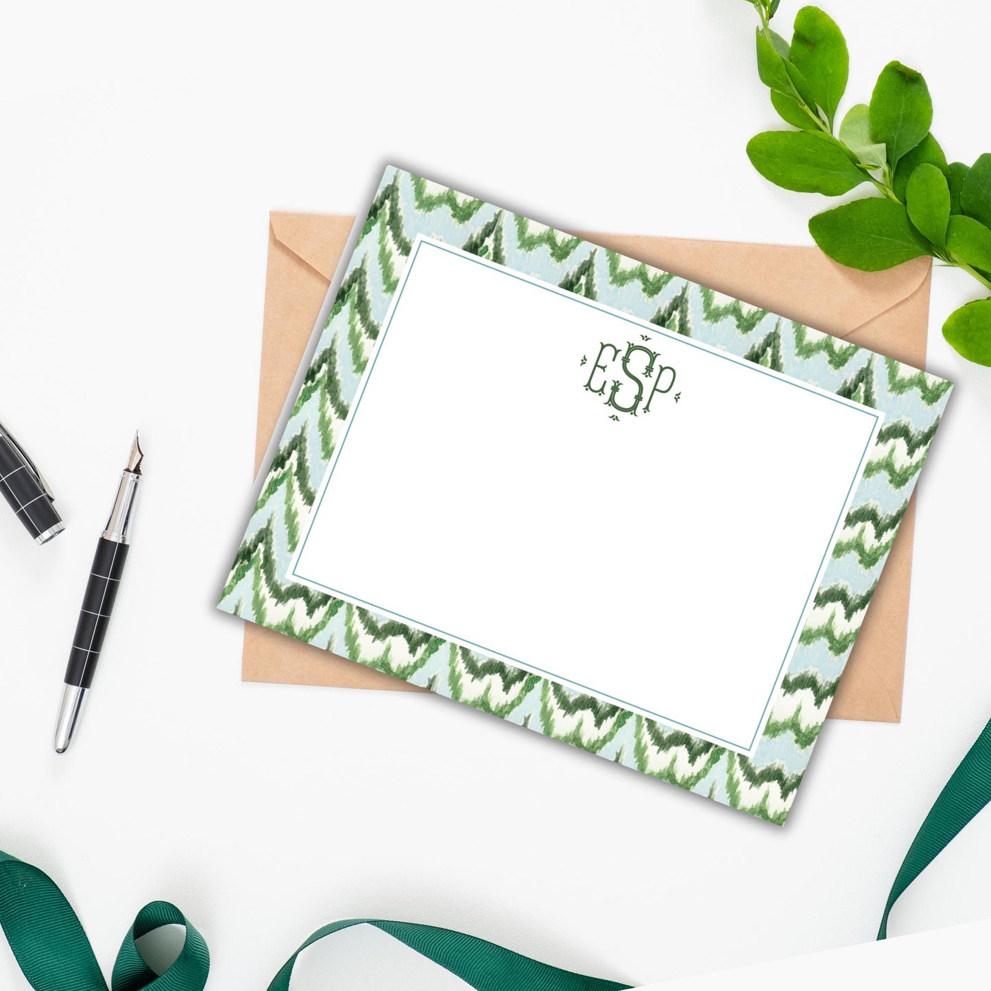 a personalized note card with a green and white zigzag pattern, placed on a white surface alongside a pen, a green ribbon, and some leaves.