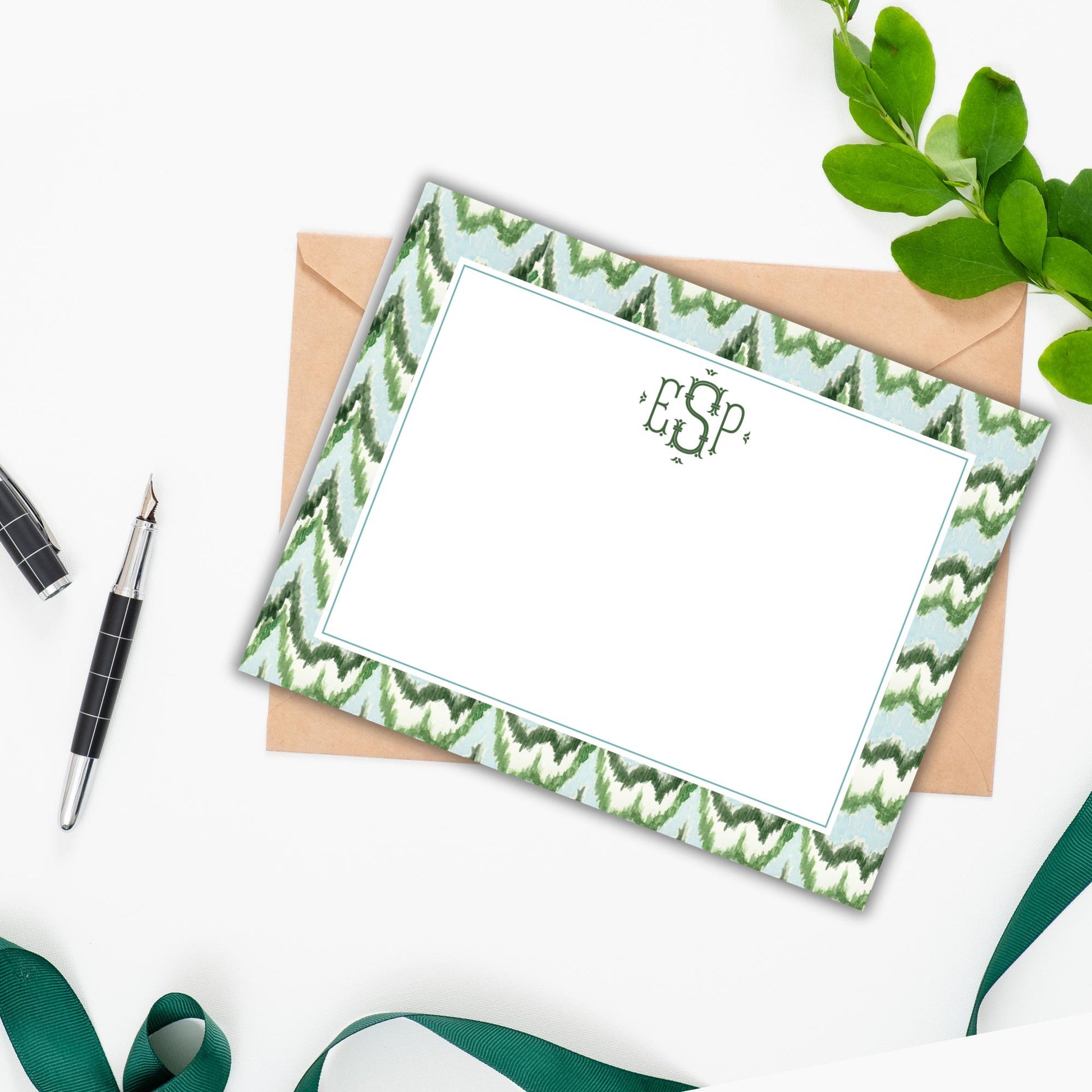 a personalized note card with a green and white zigzag pattern, placed on a white surface alongside a pen, a green ribbon, and some leaves.