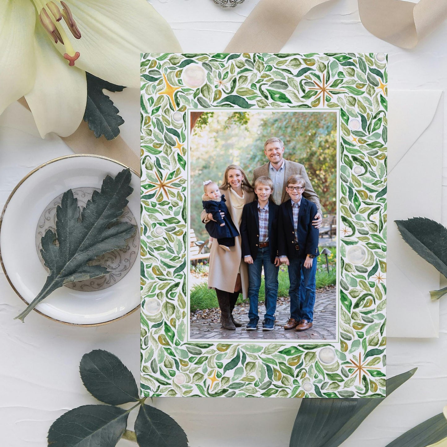 A family portrait of a group of people, including adults and children, is displayed in a frame with a green leaf pattern. The frame is placed on a white surface, surrounded by various decorative elements such as leaves and flowers.