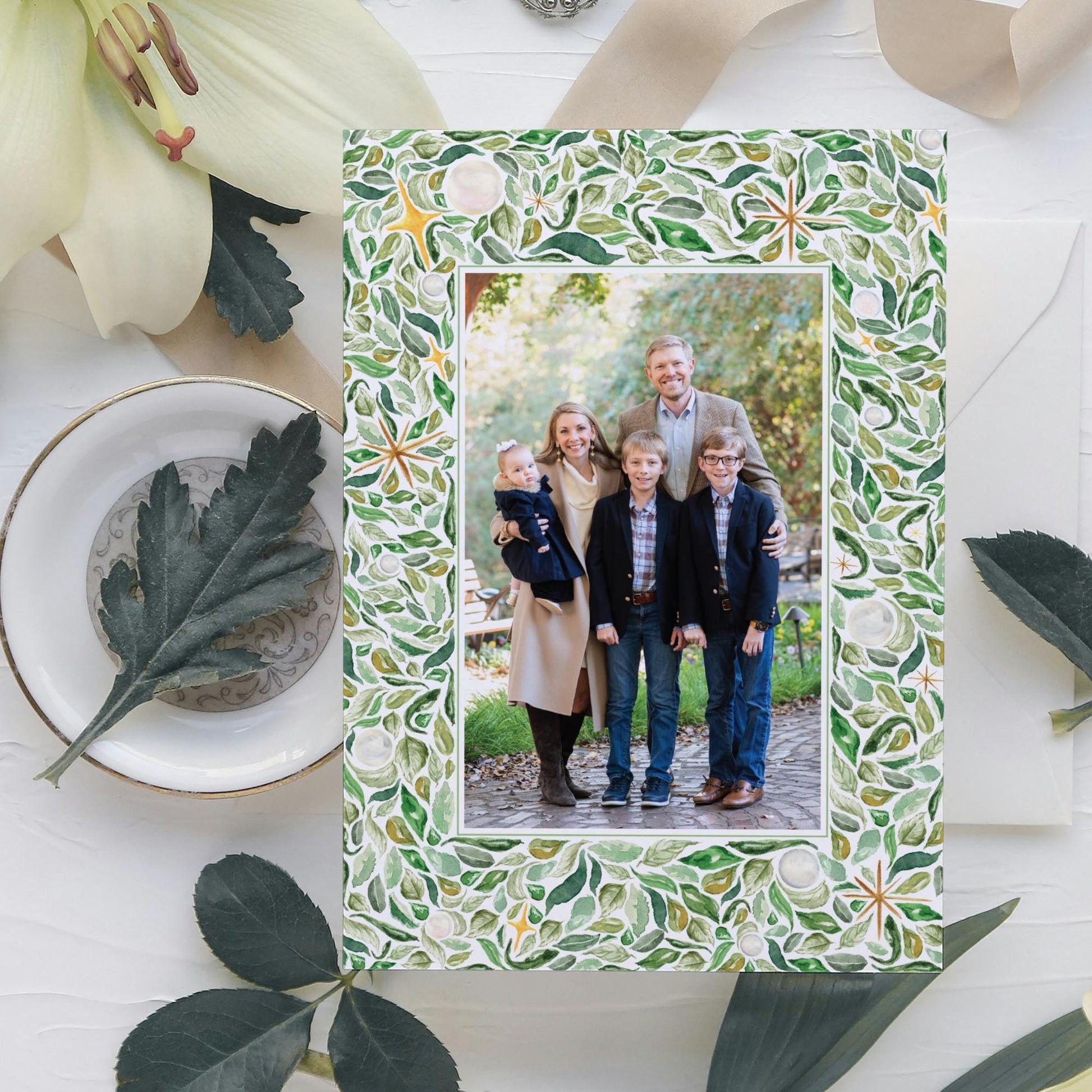 A family portrait of a group of people, including adults and children, is displayed in a frame with a green leaf pattern. The frame is placed on a white surface, surrounded by various decorative elements such as leaves and flowers.