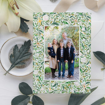 A family portrait of a group of people, including adults and children, is displayed in a frame with a green leaf pattern. The frame is placed on a white surface, surrounded by various decorative elements such as leaves and flowers.