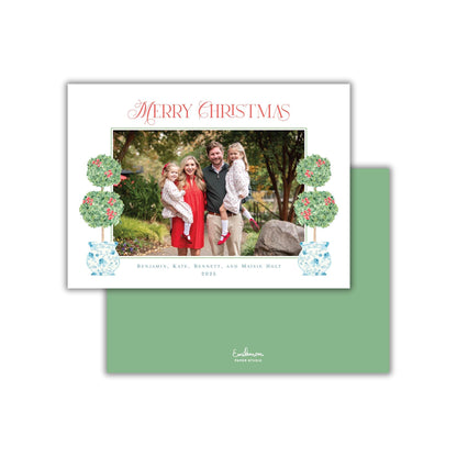 A family portrait of a man, woman, and two young girls, with the text "Merry Christmas" at the top and "Benjamin, Kate, Bennett, and Maisie Holt" at the bottom. The image is surrounded by decorative elements, including two green topiary trees and two blue and white vases.