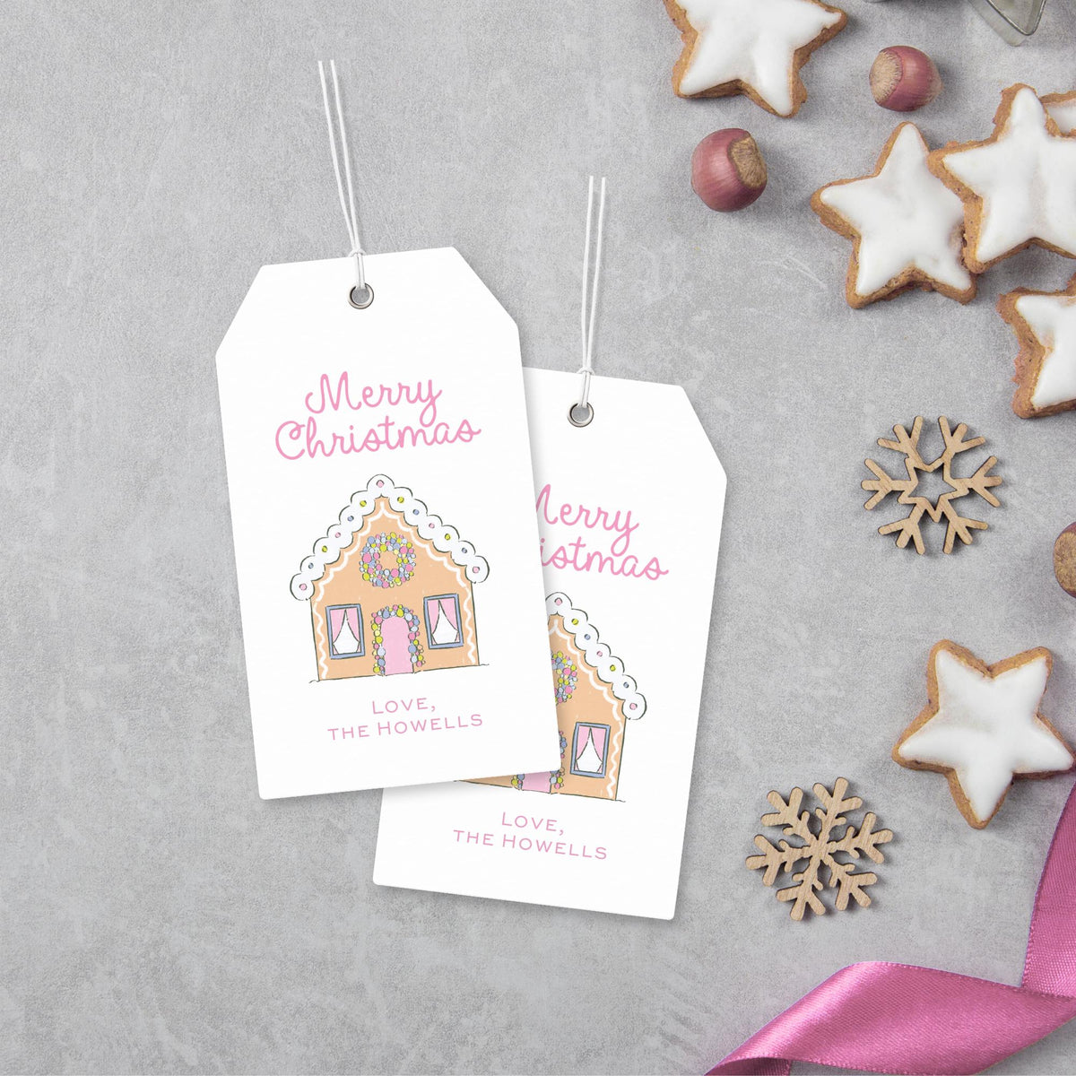 two white tags with illustrations of gingerbread houses and the text "Merry Christmas" and "Love, The Howells" printed on them, along with a pink ribbon and some cookies.
