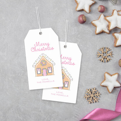 two white tags with illustrations of gingerbread houses and the text "Merry Christmas" and "Love, The Howells" printed on them, along with a pink ribbon and some cookies.