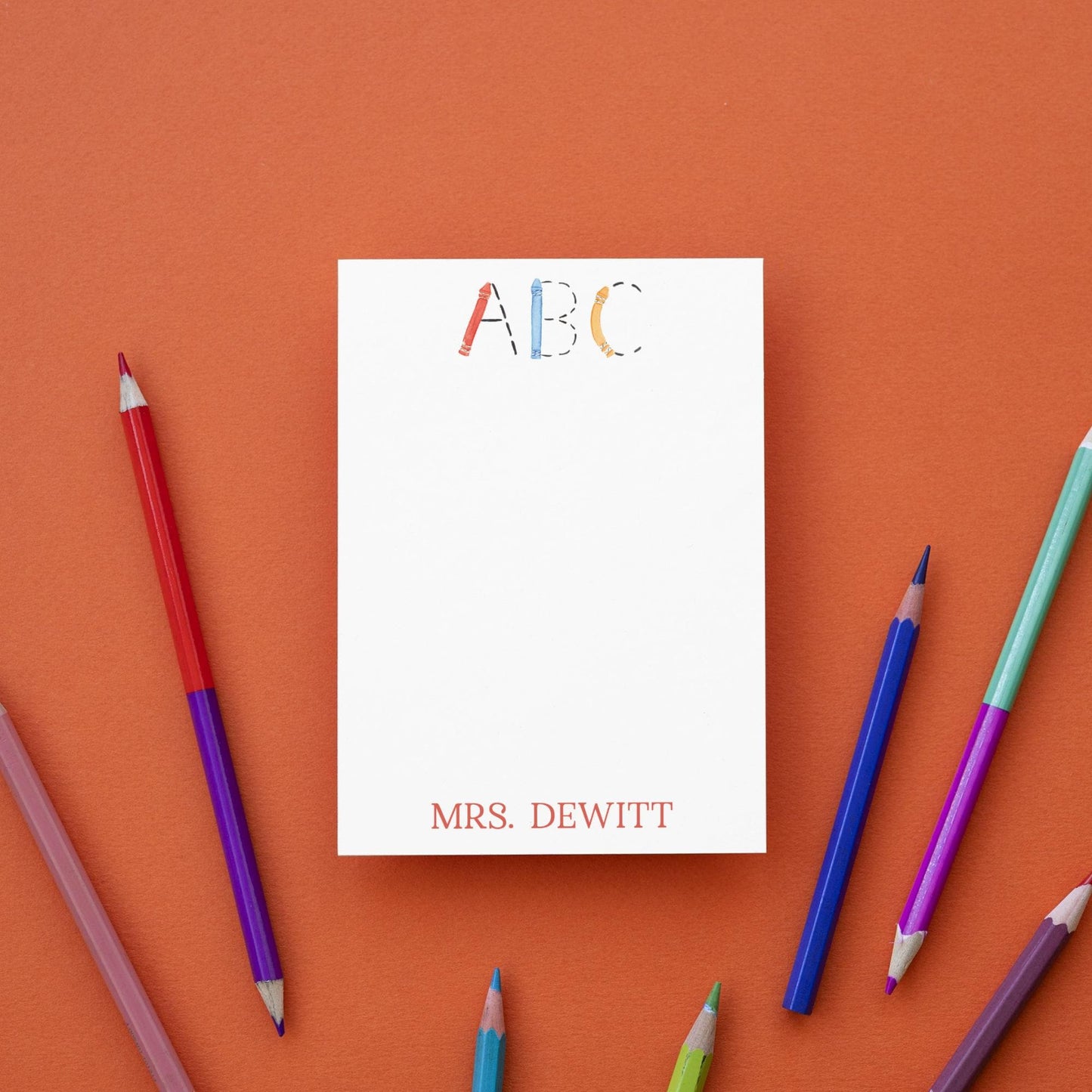 a blank white notepad with the name "Mrs. Dewitt" written on it, surrounded by a variety of colored pencils on an orange background.