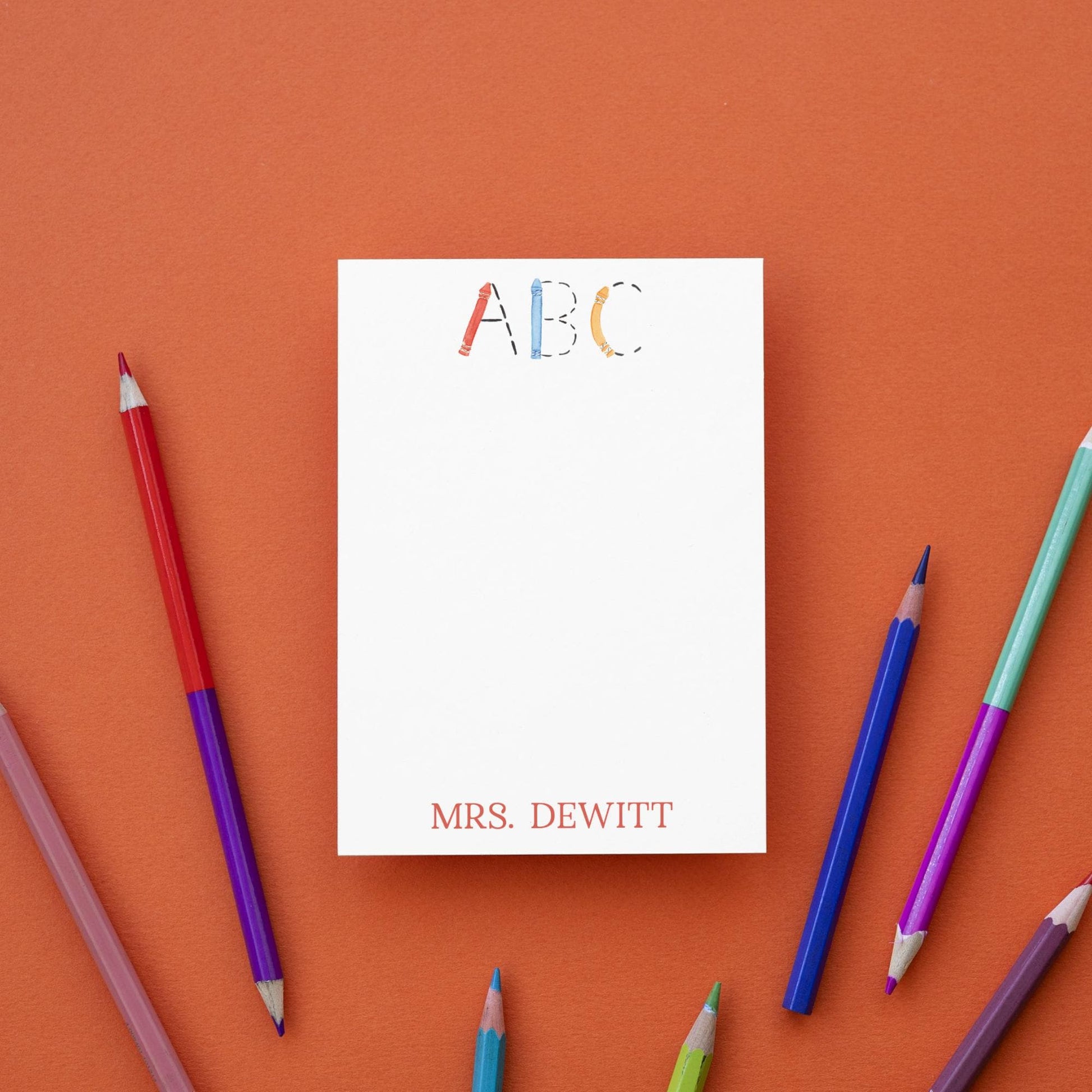 a blank white notepad with the name "Mrs. Dewitt" written on it, surrounded by a variety of colored pencils on an orange background.