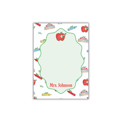 The image is a personalized nameplate with a repeating pattern of apples, crayons, and books. The nameplate has a green border and the name "Mrs. Johnson" written on it.